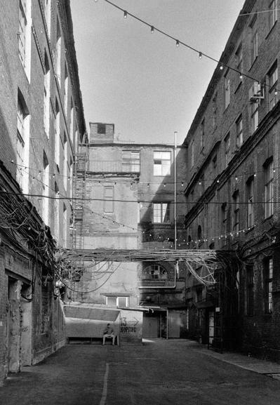 The photo shows a narrow courtyard between brick buildings in a black-and-white style. In the center of the frame, there is an aisle formed by the buildings, with electrical wires and garlands of light bulbs above it. To the left and right, there are brick buildings with windows. At the bottom of the frame, there is a part of the courtyard where a person is sitting on a bench. On the wall next to the bench, there is a sign that reads "Building 13." The overall impression of the photo is of an old industrial area or courtyard.