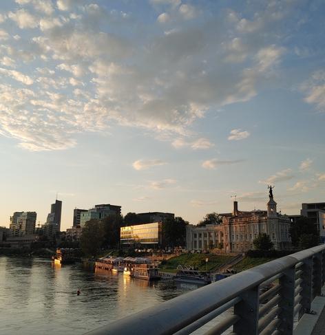 Blick von einer Brücke mit Metallgeländer, von dem man einen Ausschnitt rechts unten im Bild sieht, auf alte und moderne Häuser am Flussufer unter blauem, leichtbewölktem Himmel, die im Licht der untergehenden Sonne golden glänzen, die Silhouetten der Häuser spiegeln sich im Wasser, eine Boje ragt aus dem Wasser