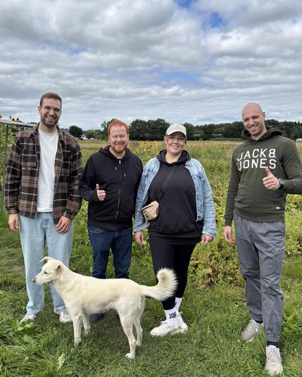 Vier lächelnde Personen stehen auf einem Feld mit grünen Pflanzen und einem leicht bewölkten Himmel im Hintergrund. Drei Männer und eine Frau posieren zusammen mit einem weißen Hund, der vor ihnen sitzt. Die Personen tragen lässige Kleidung: Der erste Mann links trägt ein kariertes Hemd über einem weißen T-Shirt und helle Hosen, der zweite Mann eine schwarze Jacke und Jeans, die Frau eine schwarze Kapuzenjacke mit einer blauen Denim-Jacke darüber, und der dritte Mann rechts trägt einen olivfarbenen Hoodie mit der Aufschrift "Jack & Jones" und graue Hosen. Die Stimmung wirkt fröhlich und entspannt.