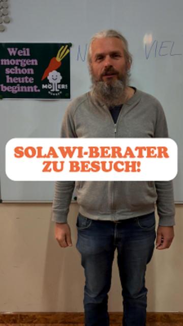 Ein Mann mit langem Bart und langen Haaren steht vor einer Whiteboard-Tafel. Er trägt eine graue Jacke und Jeans. Im Hintergrund ist ein Plakat mit der Aufschrift "Weil morgen schon heute beginnt. Möllers Gemüse" und einer Karotten-Illustration zu sehen. Im Vordergrund des Bildes steht in großen, orangefarbenen Buchstaben: SOLAWI-BERATER ZU BESUCH! Die Stimmung wirkt informativ und freundlich.