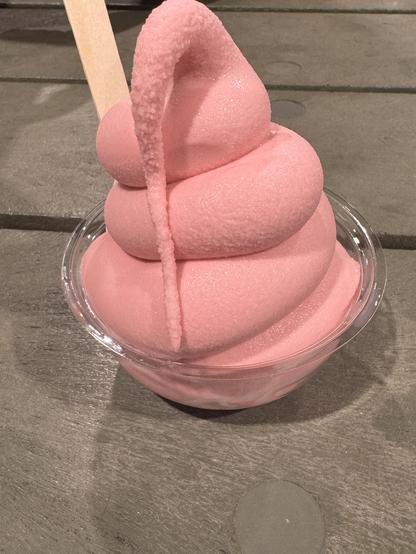 My strawberry flavoured vegan soft serve sitting in a little plastic cup on an indoor picnic table. There is a wooden spoon sticking out of the delightfully pink soft serve.