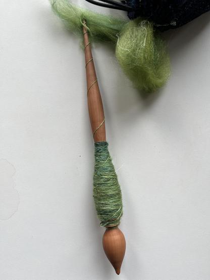 Russian style hand spindle with a metal tip, a bulb shape at the bottom and a tapered shaft. It has a fine green single on it and at the top you see the floof that it is spun from. The cob of thread starts just above the bulb shape. It's not so tidy.