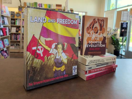 A photo taken inside Firestorm Books with the sales counter in the foreground and bookcases in the background. A boxed copy of "Land and Freedom" sits alongside a small stack of books about the Spanish Revolution, including "Lessons of the Spanish Revolution: 1936-1939" by Vernon Richards, "Collectives in the Spanish Revolution" by Gaston Leval, and "Free Women of Spain" by Martha Ackelsberg. The packaging for the game features an illustration of a working class woman in overalls waving the flag of the Second Spanish Republic flanked by the hammer and sickle on one side and the syndicalist flag on the other.