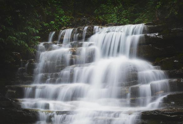 Tiered waterfall cascading over dark rock ledges in silky white streams, surrounded by lush green foliage and shaded forest.