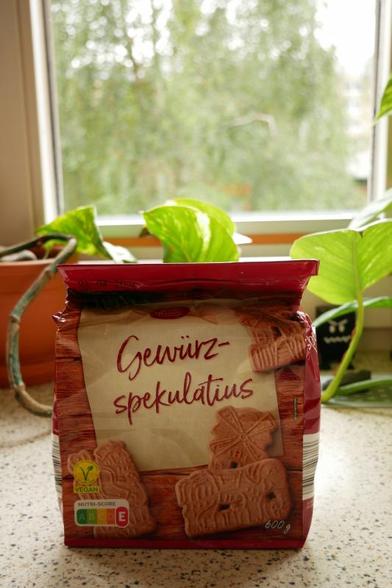 Eine Tüte Gewürzspekulatius im Vordergrund.
Im Hintergrund ist durch ein Fenster eine Birke mit sommerlich grünen Blättern zu sehen.
