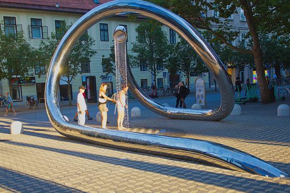 The image shows a large, modern art installation in a paved public square. The sculpture is a giant, highly polished chrome tube, twisting in a fluid, abstract loop that rises high into the air. At its apex, a showerhead sprays a continuous stream of water downwards, creating a sparkling curtain of droplets. Two people stand directly under the spray, one with their hands outstretched, seemingly enjoying the cool water on a warm day. The sculpture's reflective surface catches the low afternoon sunlight, creating bright, glinting highlights. In the background, there are trees, bikes, and historic buildings, indicating an urban setting. The ground is a light-coloured brick pattern.