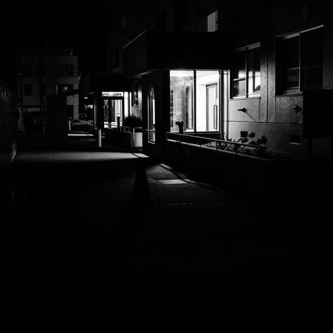 This black-and-white photograph shows a residential building complex at night with multiple illuminated entrances and walkways. The scene features modern apartment or housing buildings with clean geometric lines and appears to be a planned residential development. Several brightly lit entrances with glass doors create bright rectangular areas against the dark building facades. The buildings have multiple stories with various windows, some showing interior lighting. In the foreground, there are concrete walkways and what appears to be landscaping elements including small plants or shrubs. Metal railings are visible along the walkways and balconies. The lighting creates strong contrasts between the illuminated entrance areas and the darker sections of the buildings, emphasizing the residential complex's organized layout and accessibility features.