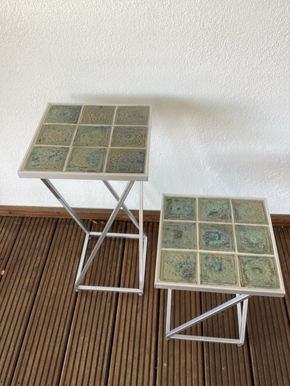 Zwei quadratische silberne Tische in unterschiedlicher Höhe mit blau, grün, grau gemusterten Fliesen.