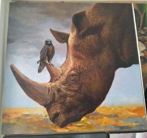 a painting of the side of a rhino's head, his two large horns sticking out, a small blue and brown hawk or falcon type bird is resting on his shorter horn, a grey cloud background, the earth gold colored
