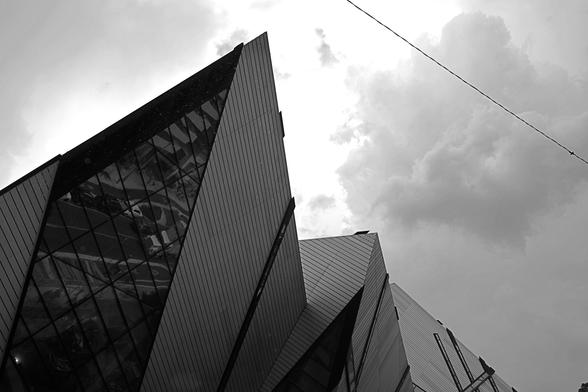 Looking up at the sharp, triangular shapes of the Michael Lee-Chin Crystal at left of the image. Beyond the structure, in the upper right, the sky is veiled in soft rounded clouds.