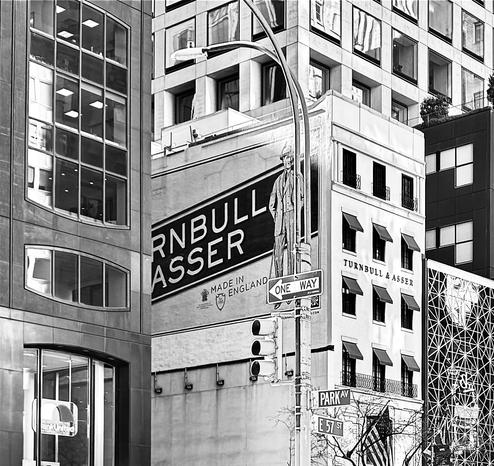 Black and white photo of the buildings on the corner of 57th St. and Park Ave., Manhattan. On the left side is a black Office building with big Modern windows. Right next to that is Turnbull and Asser, an expensive men's clothing store. The side of the building has part of the name running across it and the front of the building has small windows with neat little awnings over each one. The building next to that on the left can only be seen partially and has a large window with white lines making a geodesic pattern. Behind those are other large Office buildings