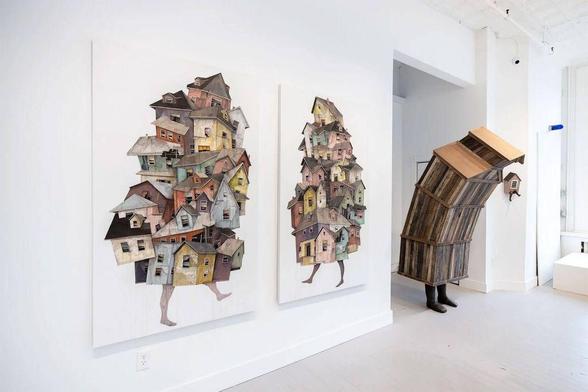 Two collages of colorful stacked houses with legs stand alongside a whimsical wooden figure resembling a building in an art gallery.