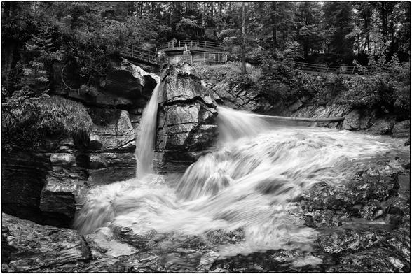 Ein kraftvoller Wasserfall stürzt in den felsigen Hexenkessel der Griessschlucht. Das schwarz-weisse Foto fängt die ungestüme Kraft des Wassers in einer weichen, langbelichteten Strömung ein, die im Kontrast zu den rauen, dunklen Felsen steht. Oberhalb des Wasserfalls sind die Strasse und der Wanderweg auf die Griesalp zu erkennen.