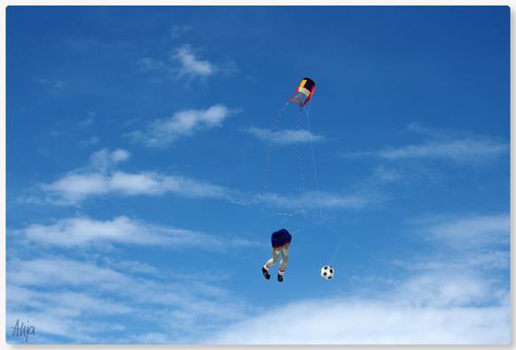 Strahlend blauer Himmel, kleine weiße Schleierwolken und Flugdrachen, einer in Form von Hosen mit Schuhen die nach einem Fußball zielen ... der andere ein buntes Ding...

Bright blue sky, small white veil clouds and kites, one in the shape of trousers with shoes aiming at a football... the other a colourful thing...