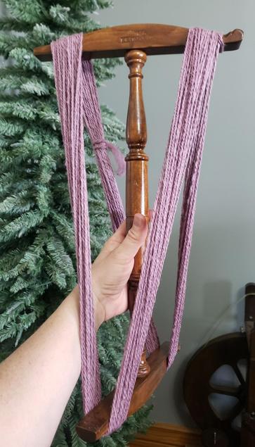 My hand holding out a niddy noddy with 3-ply dusty rose-colored yarn on it.