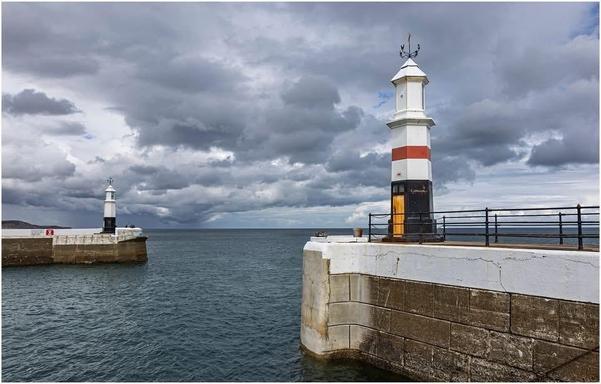 A coastal scene featuring two lighthouses on a harbour entrance. The sky is overcast with gray clouds, and the water appears calm. The lighthouses are prominently marked with red and white stripes, and a fence runs alongside the approach to the lighthouse.