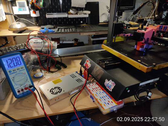 Photro of my tinkering desk. A multimeter is connected to an exposed power supply powering the working printer. It shows 12.08V.