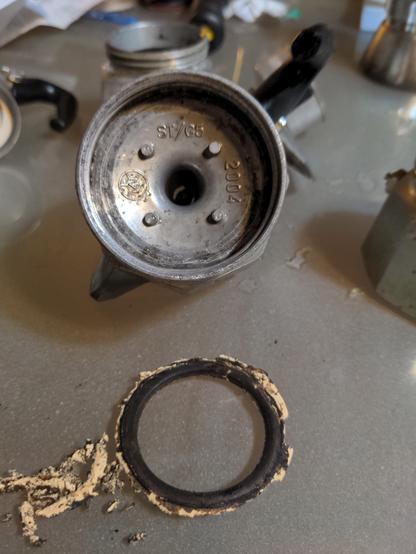 Removal of old dirty gasket and cleaning of espresso maker.  The inside and now cleaner area of the espresso maker, where the gasket was, is shown.  Dirty gasket is beside the now clean espresso maker.