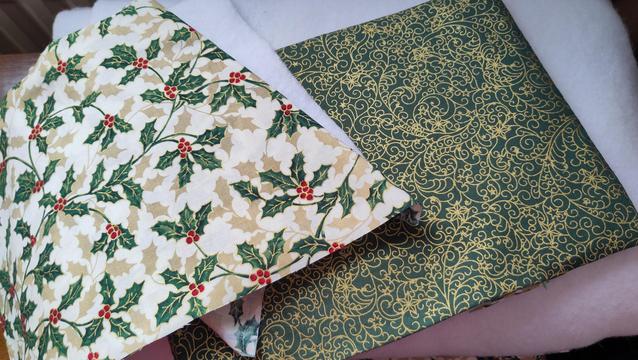 Two placemat sized pieces of fabric, one cream with green and gold holly leaves, the other green with a gold swirling pattern, on top of white padding sheeting.