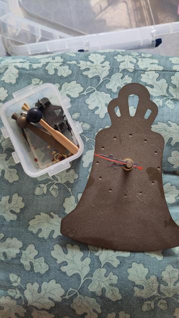 A very dusty bell shape cut out of bronze colour metal with an analogue clock face. There is a pot next to it with various bits and pieces including a brass clapper and a wooden clapoer with a blue plastic bead as the clapper ball.