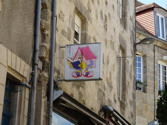 Une enseigne représentant une maison rose sous forme de personnage. Elle a un toit rose plus foncé des bras bleus avec des mains-gants jaunes et des jambes bleues avec des chaussures rose pâle et bleues. Elle tient un livre bleu foncé qu'elle semble lire intensément avec ses gros yeux ronds façon dessin animé.