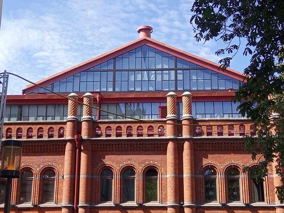 Red brick building. Windows with rounded tops are placed in groups of three with a pair of brick pillars between each group. Above it two storeys of mostly windows; the top storey is triangular due to the slanted roof. A black iron grid forms standing rectangles with glass in them. The construction lets a lot of light in to the centre of the large market hall.