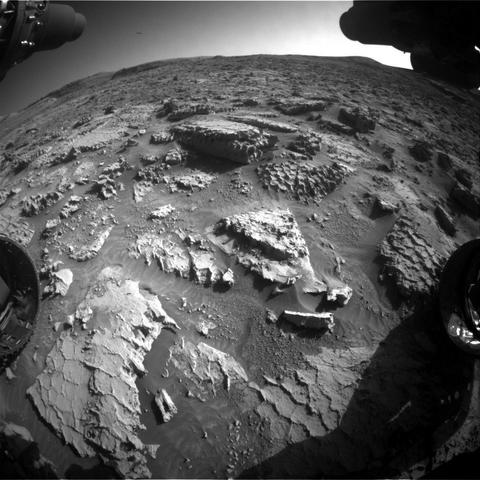 An extreme wide-angle photo of the Martian surface in front of the Curiosity rover shows extremely rocky, rough terrain, with flat rocks in the foreground showing apparent honeycomb-shaped patterns on their exposed faces, and a larger rock at the center of the image, looking like an offset stack of waffles. Portions of the rover are visible in each corner of the frame, with two wheels visible, but shadowed, in the lower left and right corners.
