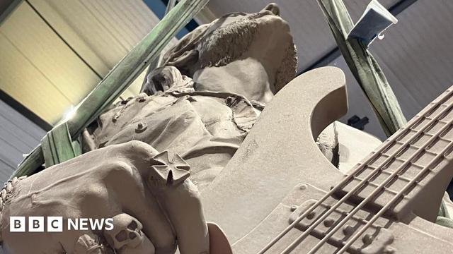A statue of a man holding a guitar, viewed from below and looking straight up to his chin. The fingers he is using to play the guitar have rings on them, including one of a skull.