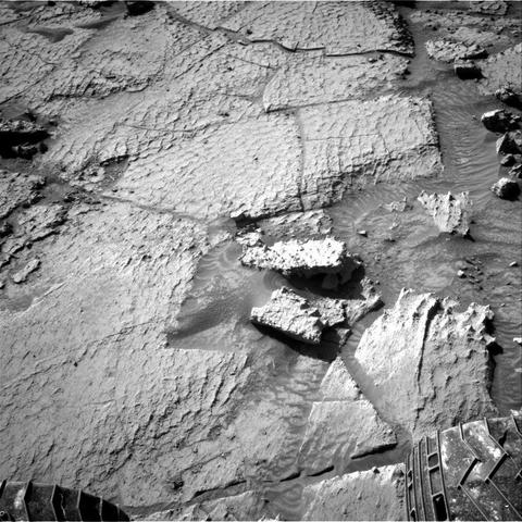 A grayscale photograph from the Martian surface showing mainly flat terrain, looking like it has fractured into various large, interlocking, angular plates. The plates appear textured, looking similar to reptile scales. At image center, two smaller rocks poke above the surface, angled similarly to the plates, but rising above them.