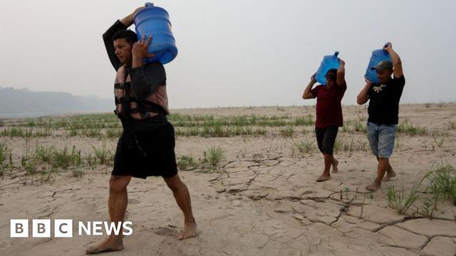 Three men carry drinking water above their heads in large blue plastic tanks along a dry sandbank of the Madeira River in northern Brazil