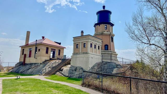 A historic lighthouse and adjoining brick building sit on a rocky hill under a blue sky, with a grassy pathway and a few trees in the foreground.