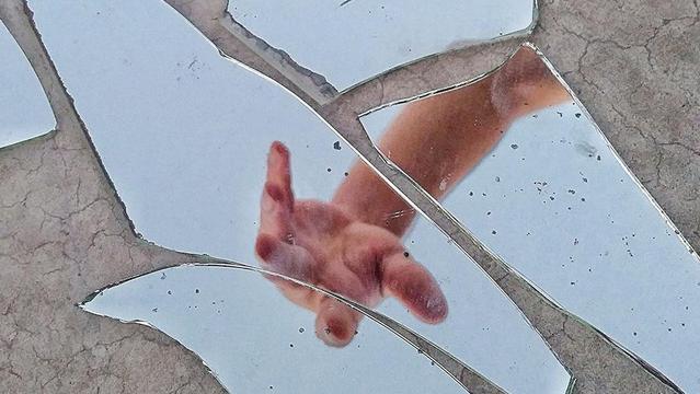 A hand reaching upward is reflected in several broken pieces of a mirror scattered on a cracked, dry surface.