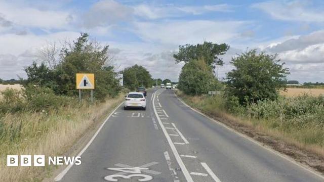 A road bends slightly to the right, there are white road markings on the side and centre of the road surface. Two vehicles are ahead travelling in the left hand lane. Bushes and grass lines both sides of the road. There is a sign on the left warning of an upcoming right hand turn.