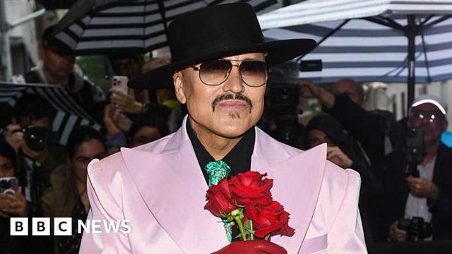 Willy Chavarria on a red carpet with photographers all around wearing a black hat, sunglasses and a pale pink tuxedo with exaggeratedly large lapels and holding three red roses