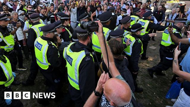 Police officers detain a protester during a rally aimed at challenging the government's proscription of Palestine Action under anti-terrorism laws, in in London, Britain on 9 August. The picture shows a cluster of police in high-vis jackets, holding a man, with a crowd surrounding them. In the foreground, a man takes pictures with a camera.
