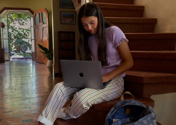 A young woman with long dark hair sits on wooden stairs wearing a light purple ribbed top and striped pants, working on a space gray Apple MacBook Air with M1 chip. She has a blue patterned backpack beside her, and the warm-toned home interior features tile flooring, framed artwork on the walls, and an open doorway with greenery visible outside.