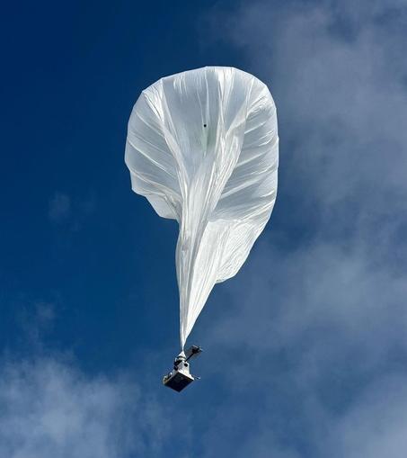 A hot-air balloon hoisting a complex radar instrument, mid-flight.