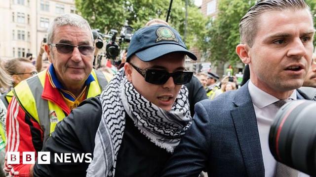 Liam Óg Ó hAnnaidh (wearing a blue keffiyeh , dark blue cap and sunglasses arrives at court. To his right is his solicitor and to his left is a man in  a hi-iz hacket