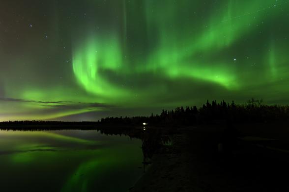 The northern lights burn in green flame across a starry night sky, Ursa Major visible on the left. Below, a still lake reflects back the sinuous curves of light on the left; along the horizon, the forest slopes up into distinct tree shapes on the right. There is a little lit building in the centre distance. 