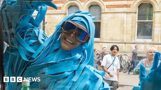 A man is dressed in an elaborate costume made of long strips of blue fabric ans is wearing large goggles. He is waving through a window at someone sitting inside. There are people walking on the street behind him