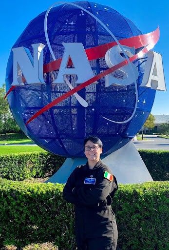 A person wearing a black NASA flight suit stands smiling with arms crossed in front of the large blue NASA globe sculpture. The Irish flag patch is visible on their sleeve, and the sky is clear and blue in the background.