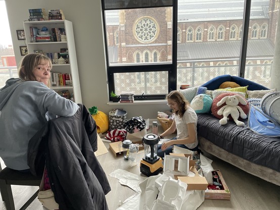 My mom watching as my wife unpacks a yellow Vitamix from the box. There is Christmas wrapping scattered about in our apartment.