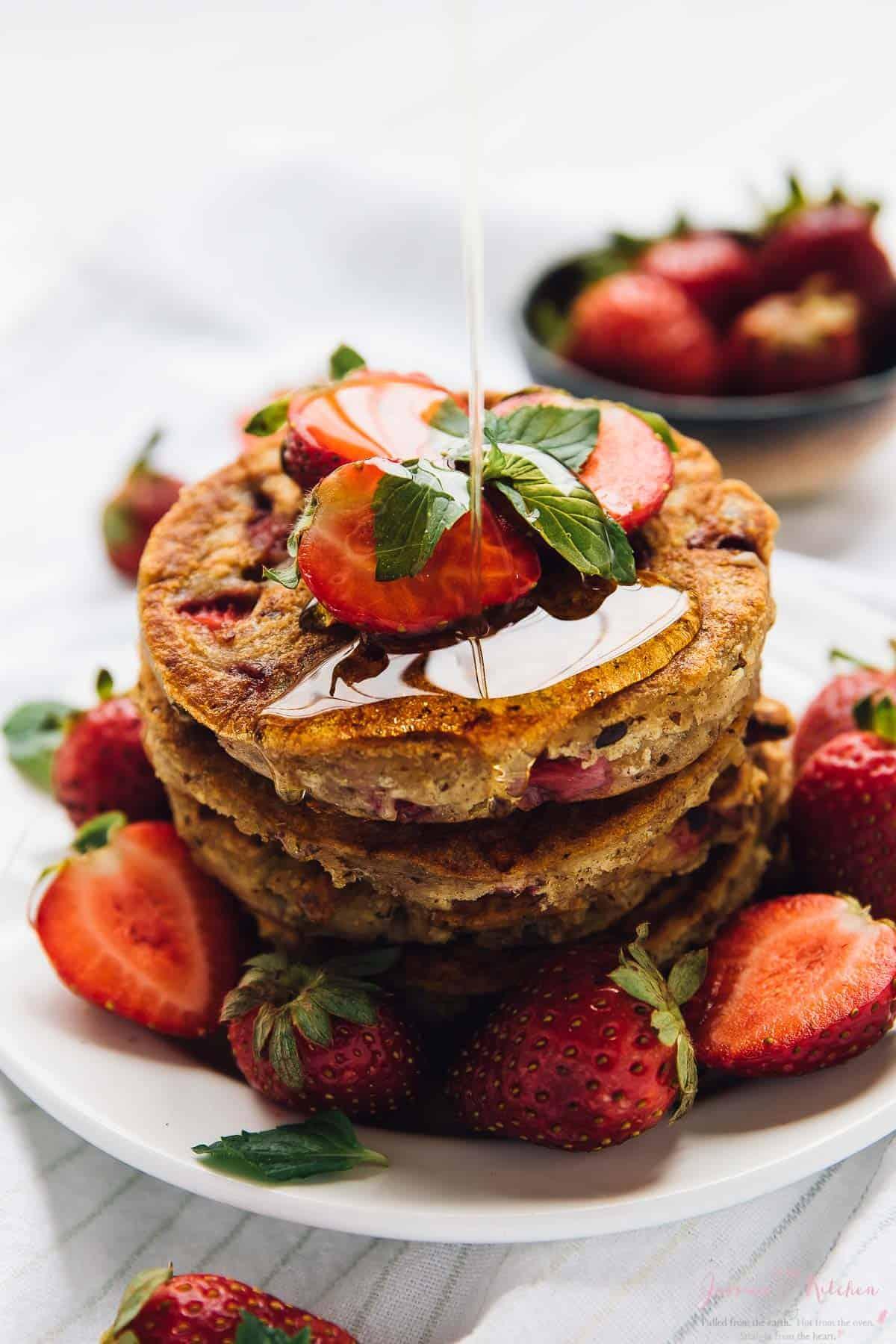 "A stack of vegan strawberry buttermilk pancakes topped with fresh strawberries, mint, and a drizzle of maple syrup, surrounded by halved strawberries on a white plate."