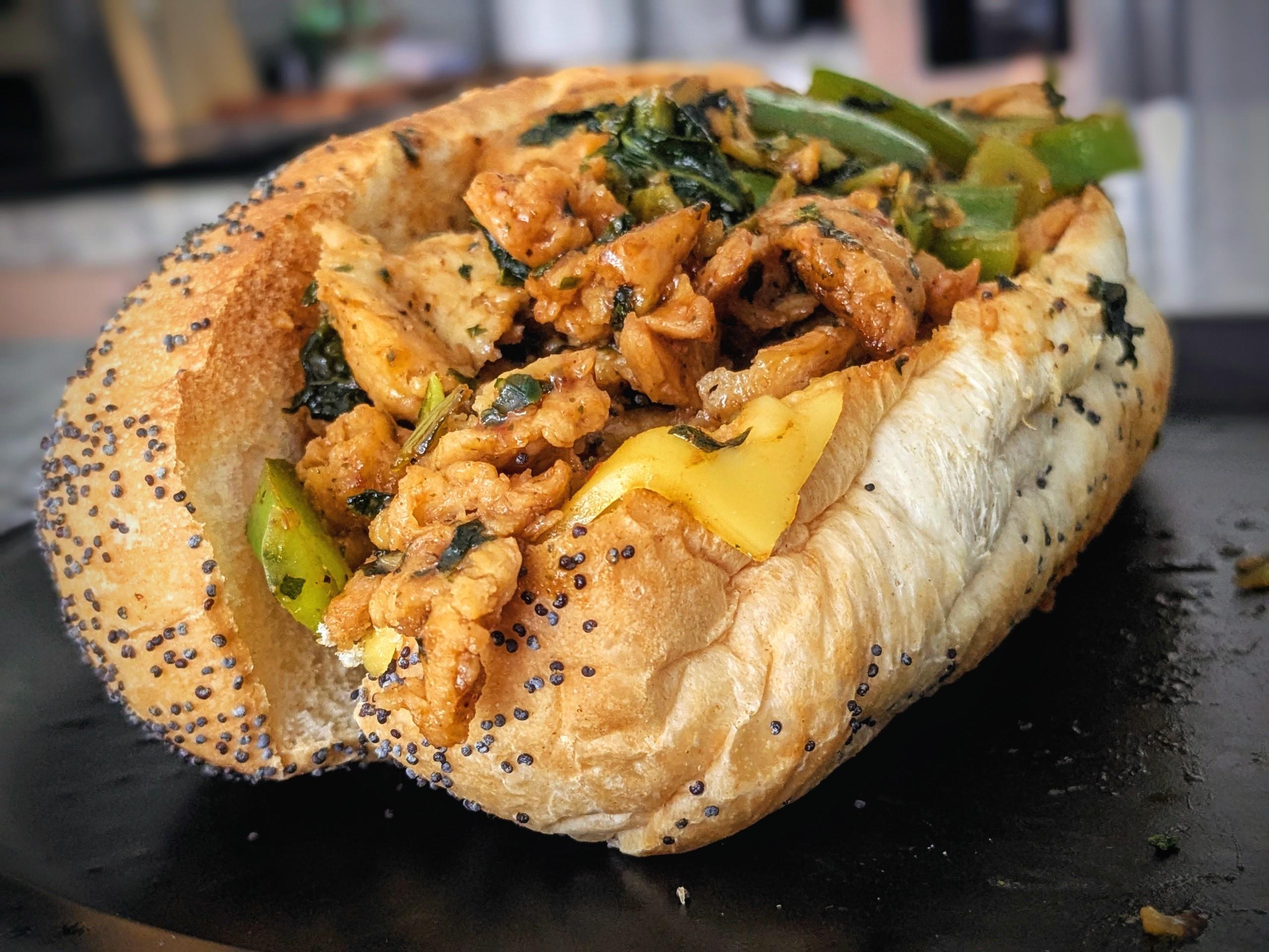 Bbq soy curls on a seeded hoagie roll, with green peppers, turnip greens, and melted cheese. On a black plate.