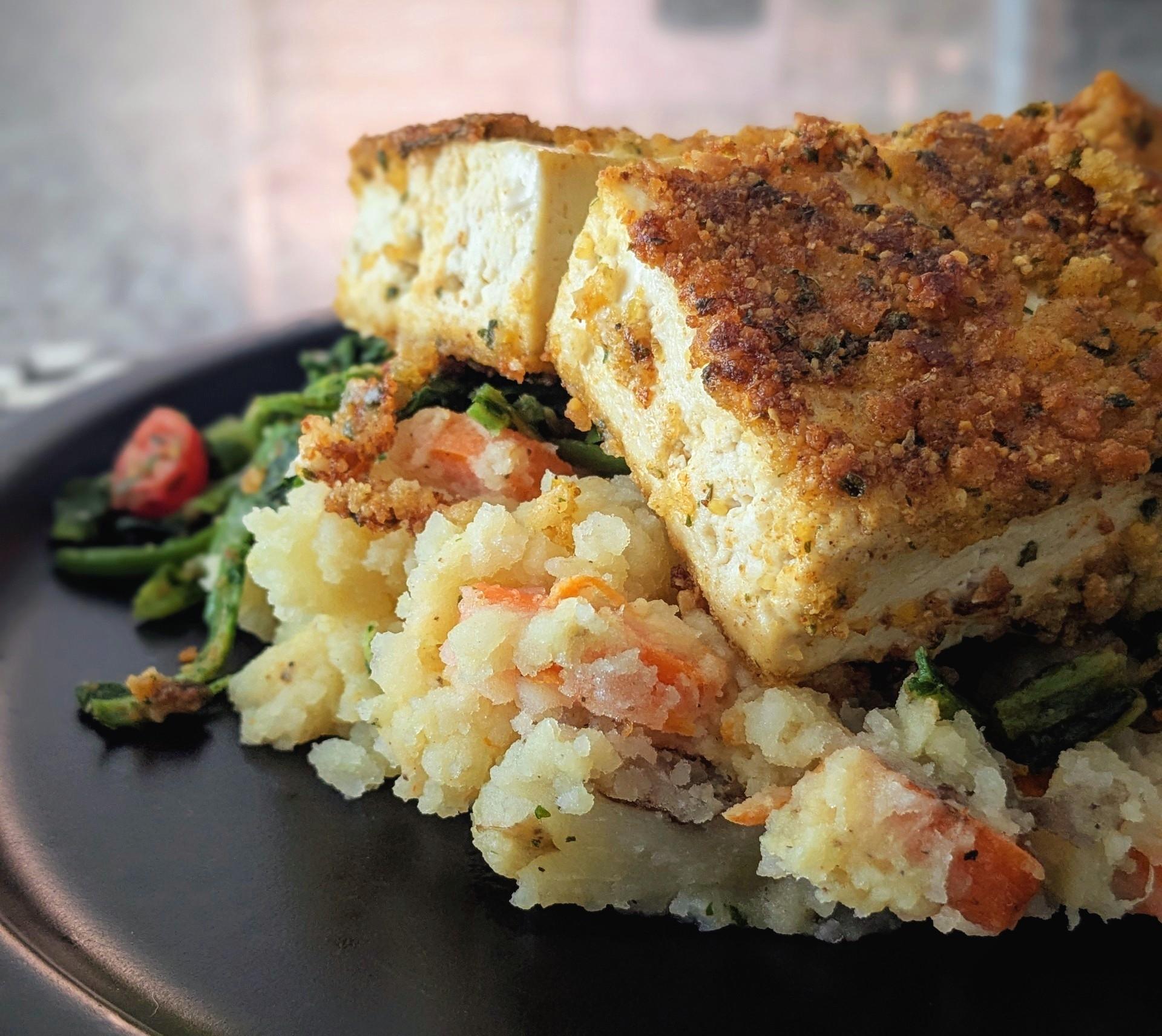 Two slices of tofu with a falafel breading crust, over mashed potatoes with carrots, and sauteed green beans and turnip green. On a black plate.