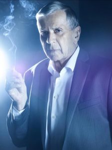 Actor William B Davis from XFiles. A blue background. he's wearing a suit and smoking a cigarette. 